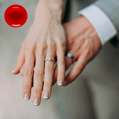 A woman's hand on top of a man's hand both wearing wedding rings with a red blood cell in the corner